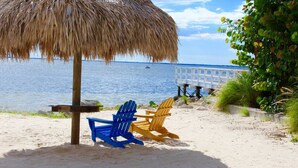 A beach with a thatched umbrella, two Adirondack chairs, and a view of the water.