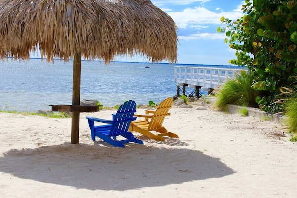 A beach with a thatched umbrella, two Adirondack chairs, and a view of the water.