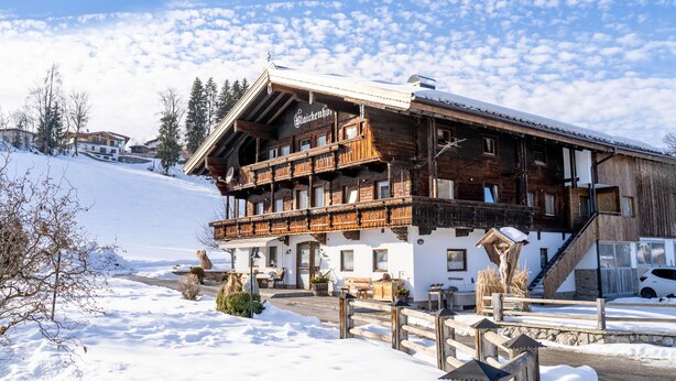 A wooden chalet with a balcony, surrounded by snow-covered ground and trees.
