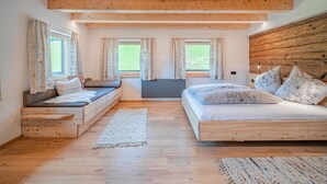 A bedroom with a wooden ceiling, two windows with curtains, a bed with patterned pillows, a bench, and a rug.