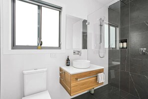 Une salle de bain moderne avec des toilettes blanches, un meuble-lavabo en bois avec un lavabo blanc, un grand miroir et une douche séparée par une paroi de verre.