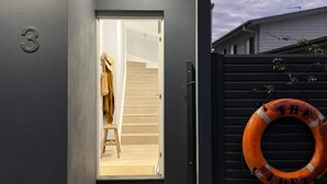 A modern building entrance with a grey facade, a number '3' sign, a wooden staircase leading inside, and a lifebuoy on a black fence. - The Loft / Luxurious 2 Bed Abode / Walk to Marina (Shell Cove)
