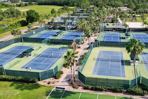 An aerial view of a tennis facility with multiple courts, surrounded by palm trees and greenery.