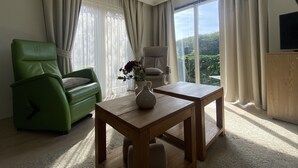 Uma sala de estar moderna com um sofá verde, uma mesa de centro de madeira e vista para áreas verdes através de grandes janelas.