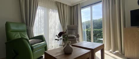 A modern living room with a green sofa, a wooden coffee table, and a view of greenery through large windows.