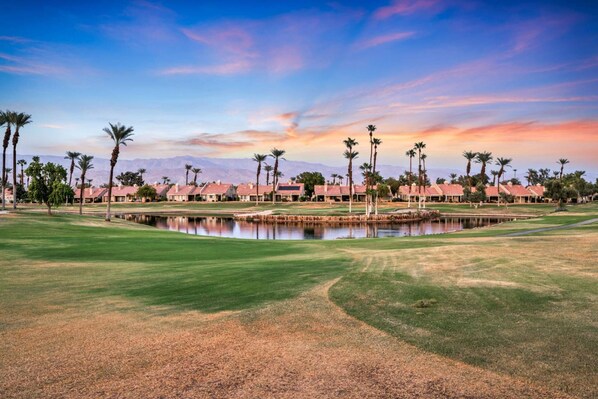 A golf course with a pond, palm trees, and a row of red-roofed buildings in the background.