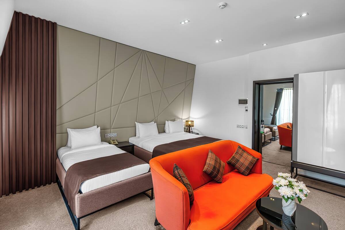 A modern hotel room with two beds, an orange sofa, a small table with a vase, and a large mirror.