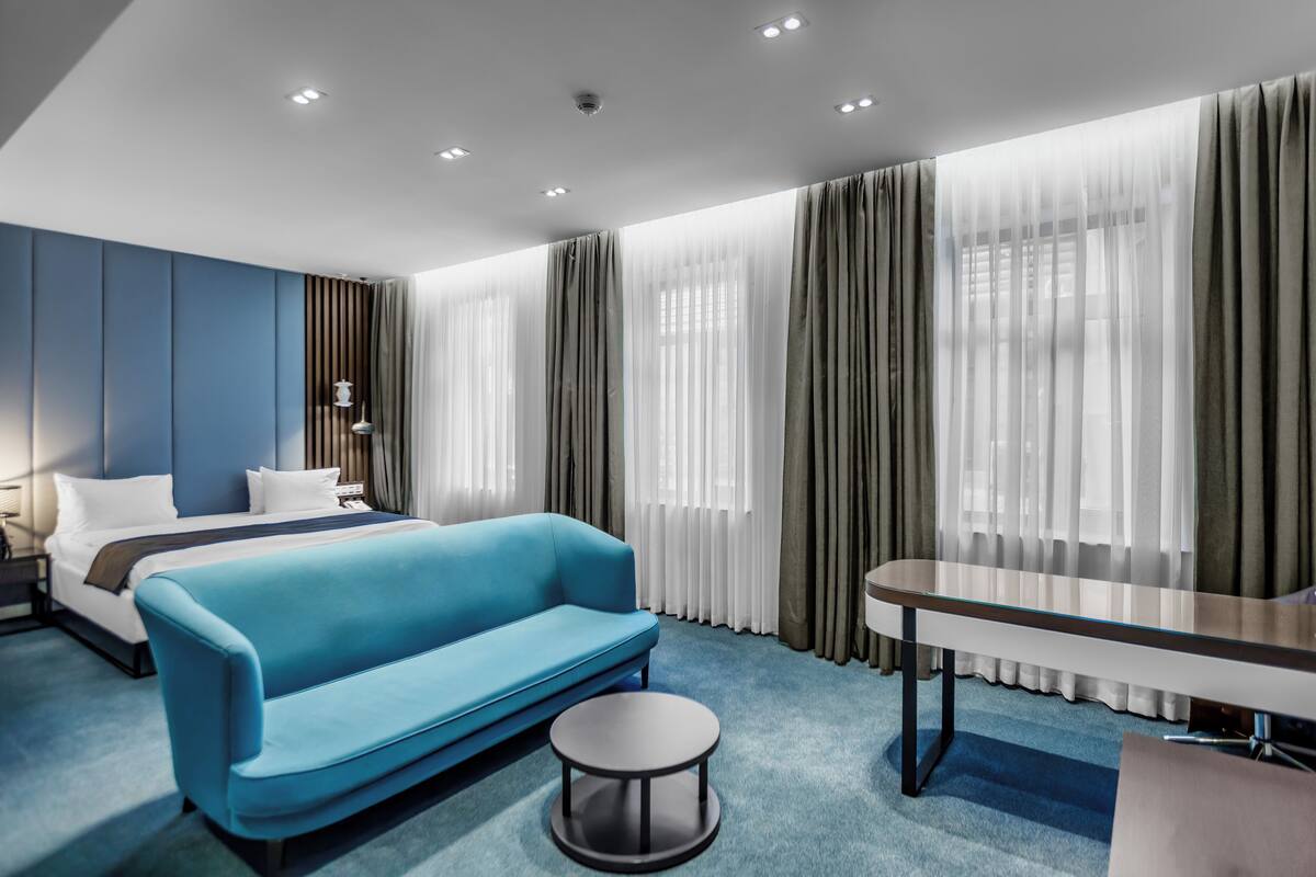 A modern hotel room with a blue sofa, a round coffee table, a bed with white linens, and a desk with a chair.