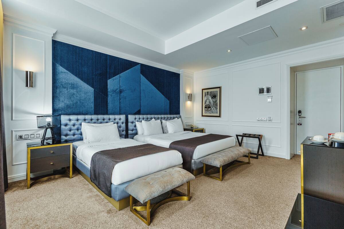 A hotel room with two beds, a large abstract painting, a bedside table with a lamp, and a bench.