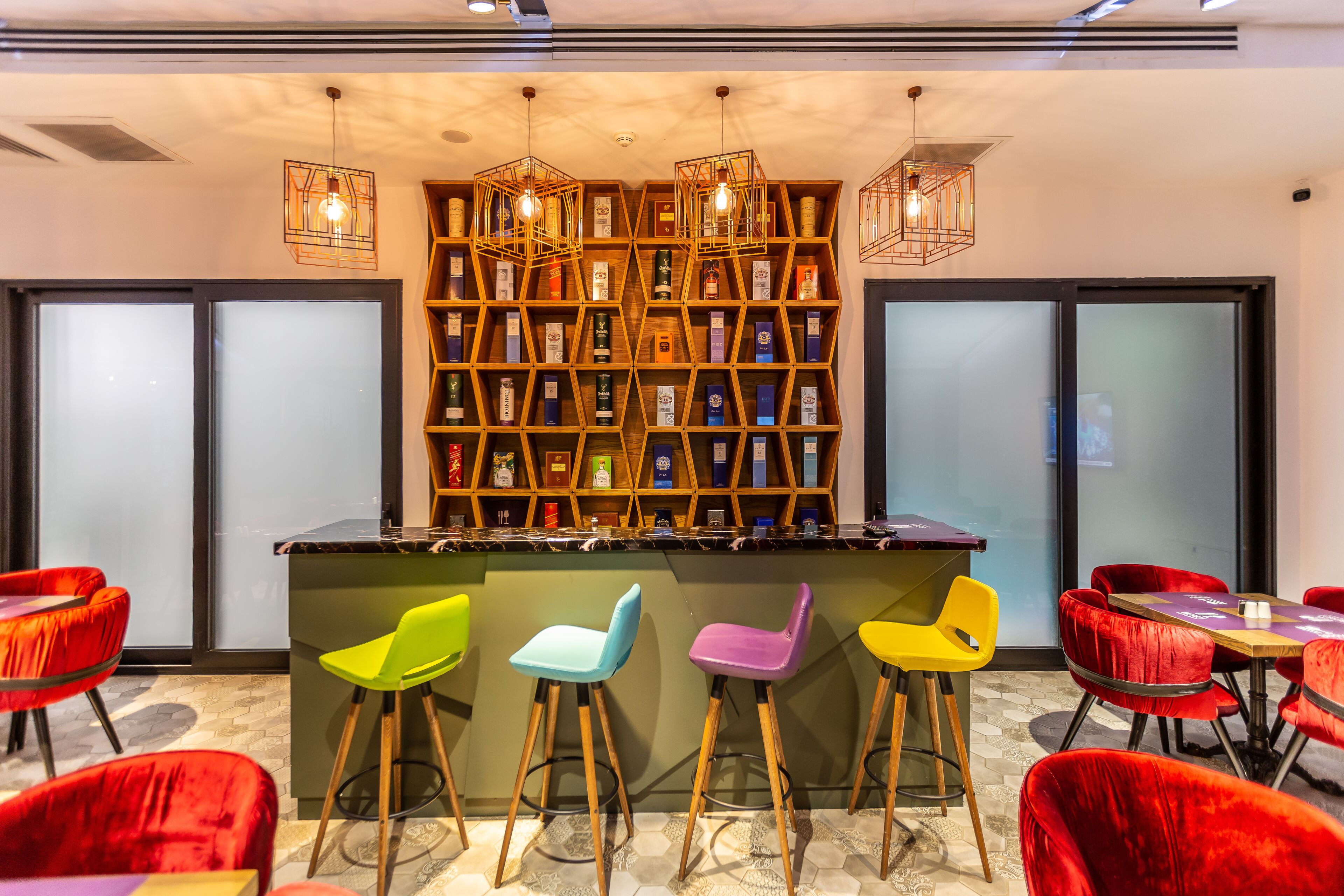 A bar area with a wooden bookshelf, colorful bar stools, and pendant lighting.