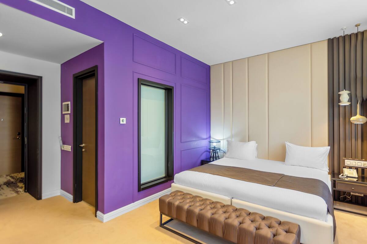 A modern hotel room with a large bed, a bench at the foot of the bed, and a window with blinds.