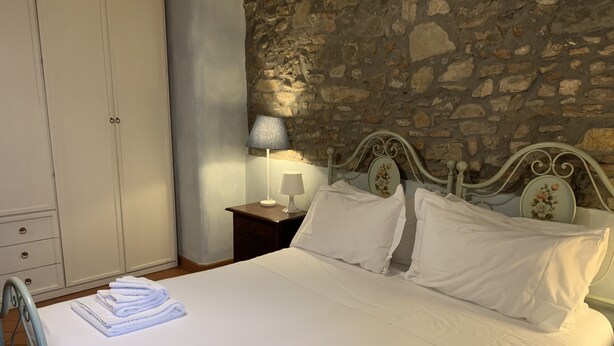 A neatly made bed with white linens, a metal-framed headboard, and a nightstand with a lamp. A wardrobe is visible to the left, and a stone wall is behind the bed.