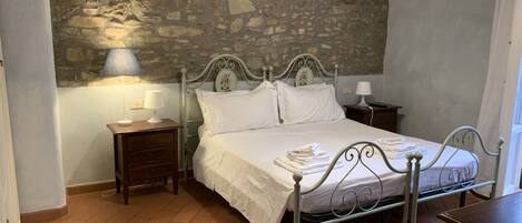 A bedroom with a stone wall, a metal bed frame, a wooden nightstand, and a bedside table with a lamp.