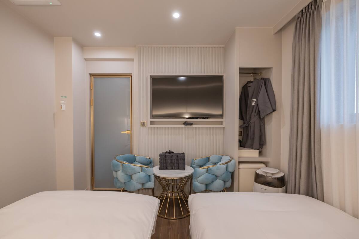 A hotel room with a bed, two armchairs, a small table, a TV, and a coat rack.