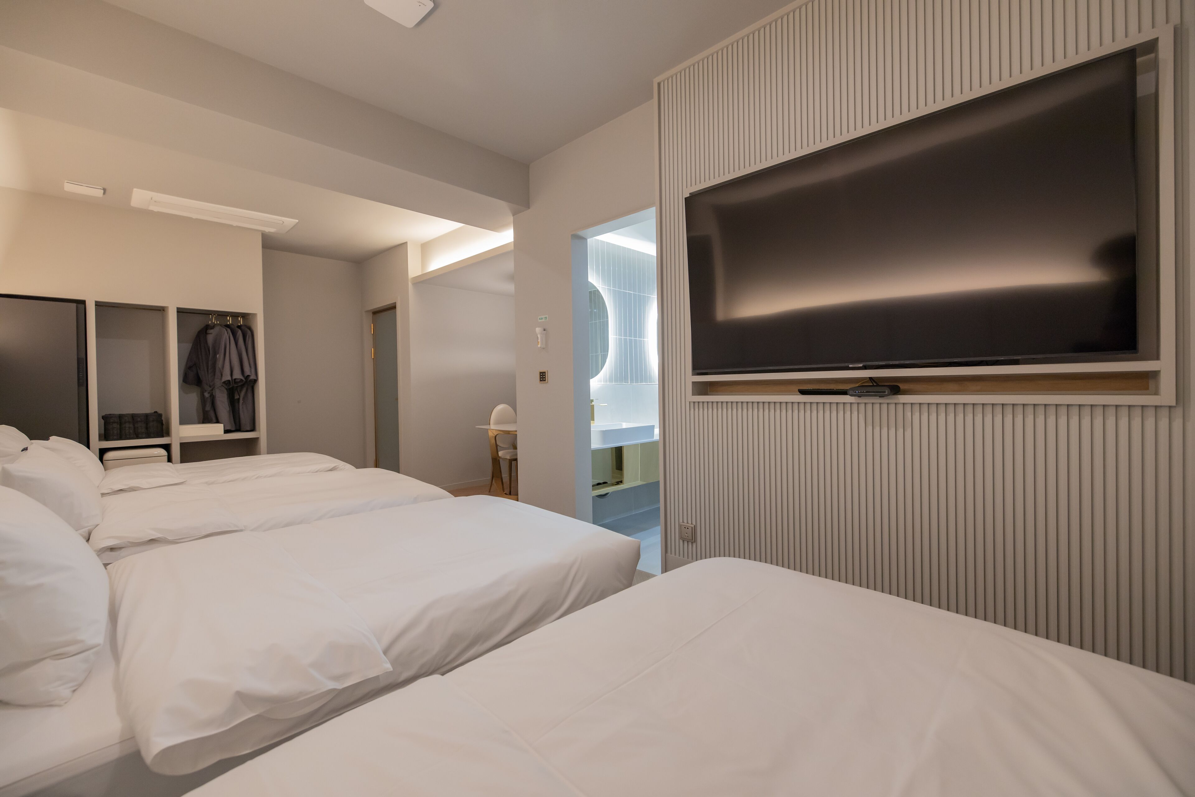 A modern hotel room with three beds, a large flat-screen TV, and a walk-in closet with hanging clothes.