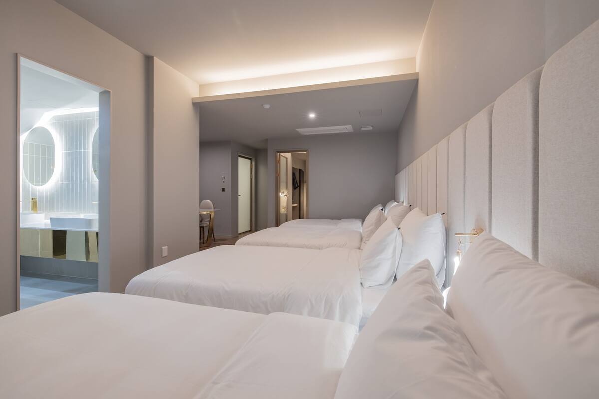 A hotel room with three beds aligned against a wall, a bathroom with a modern design, and a hallway leading to another room.