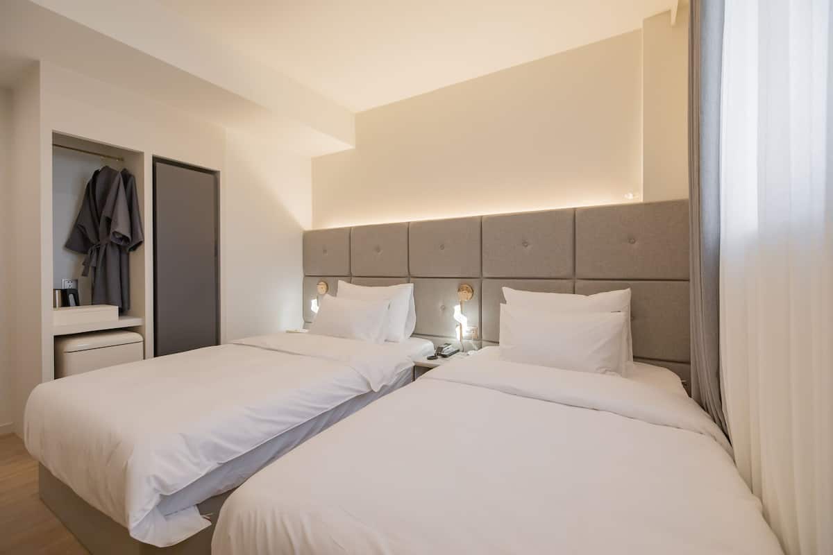 A hotel room with two beds, a wardrobe, a bedside table with a lamp, and a headboard with built-in lighting.