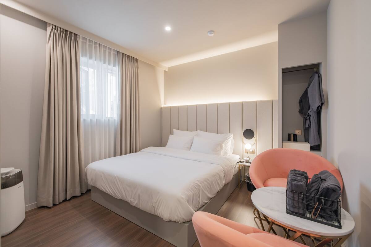 A modern hotel room with a large bed, a bedside table with a lamp, a small round table with towels, and a wardrobe.