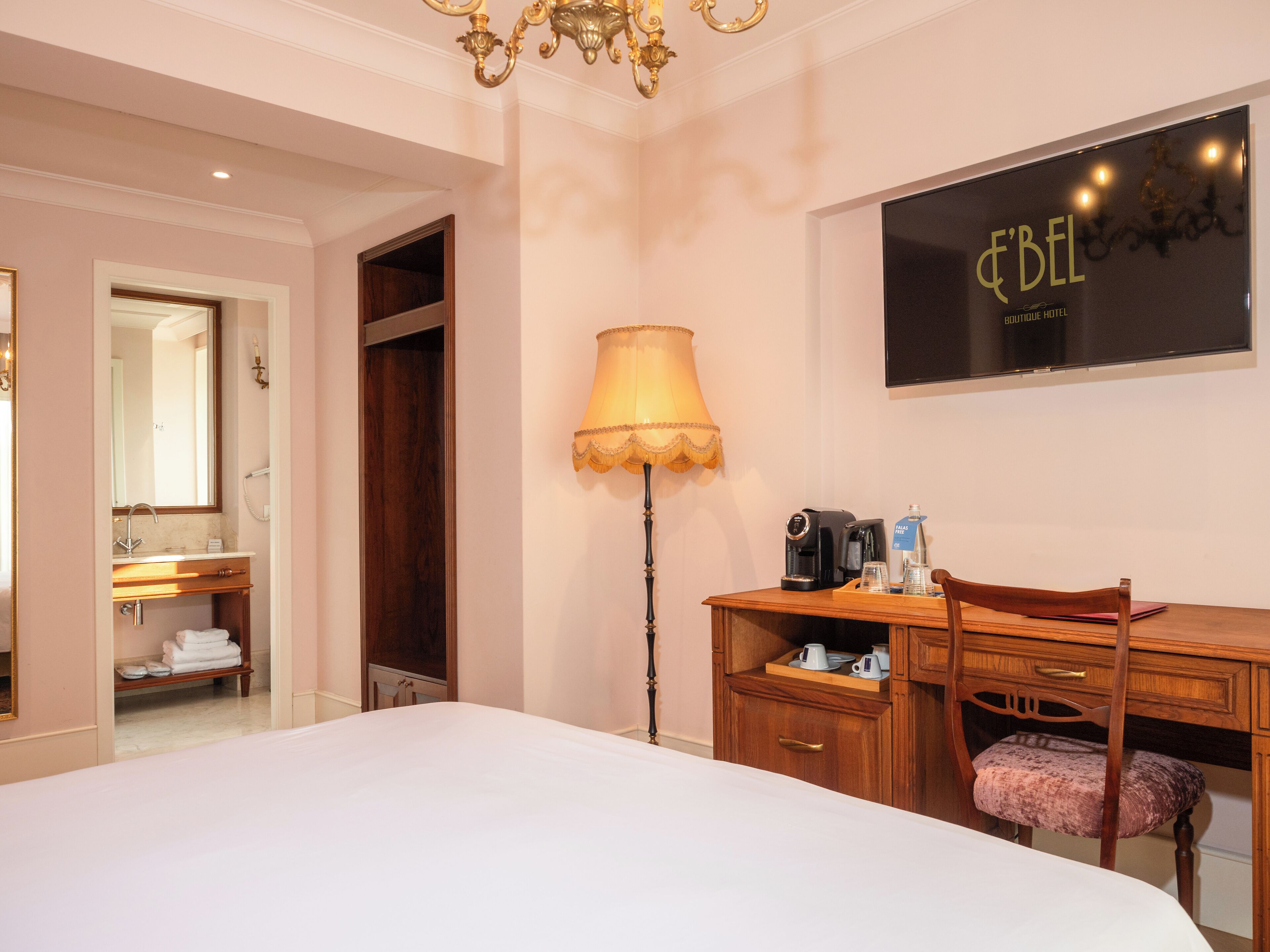 Une chambre d’hôtel comprenant un lit, un bureau avec une chaise, une lampe, une machine à café et un téléviseur affichant le nom de l’hôtel.