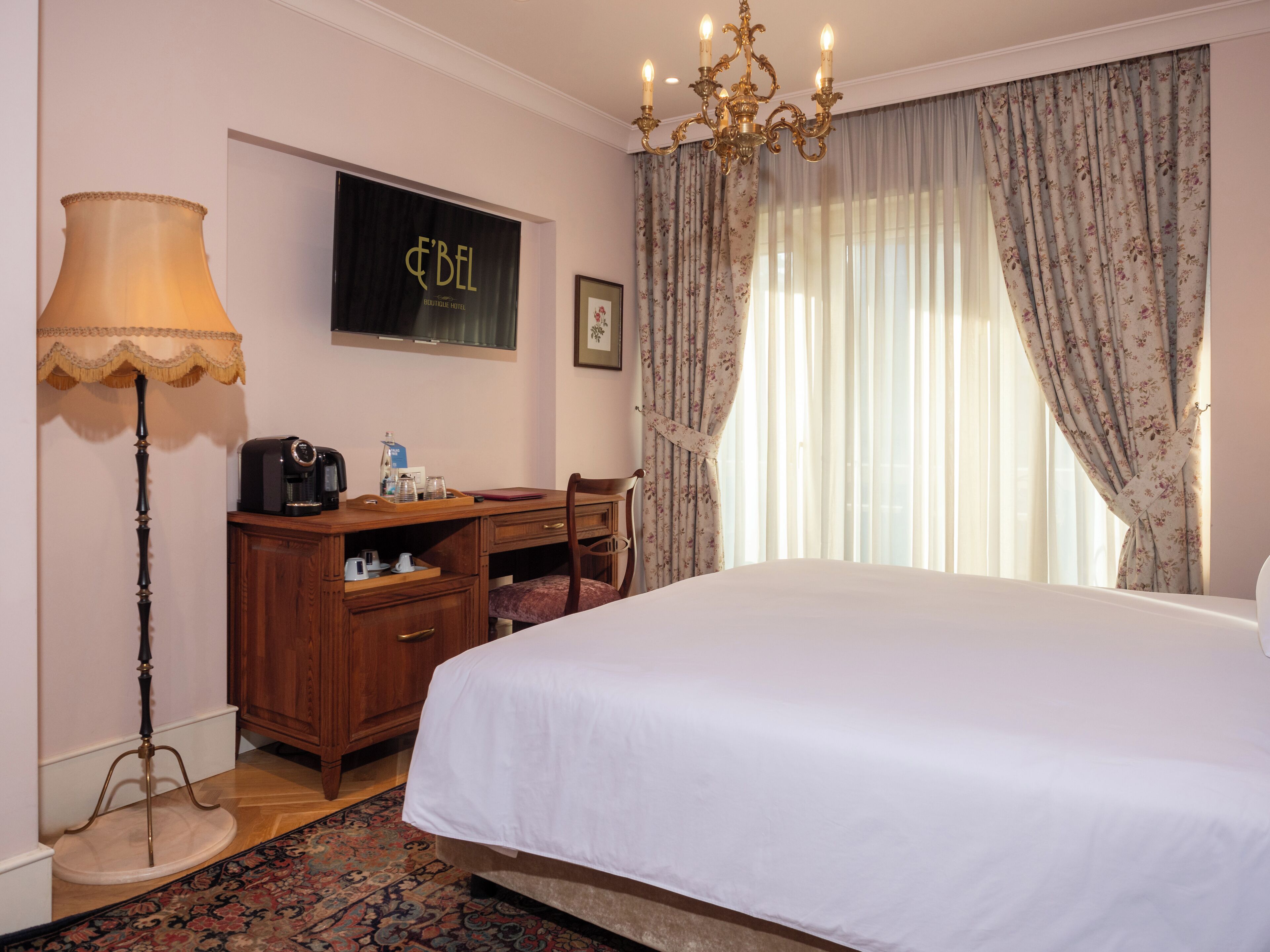 A hotel room with a bed, a desk with a lamp, a coffee maker, and a framed picture on the wall.