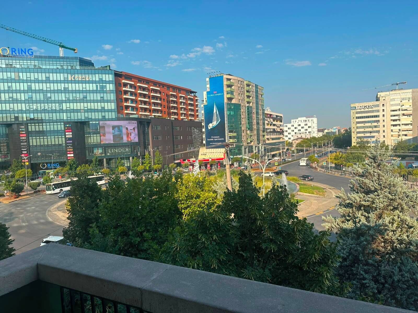 Un paisaje urbano con edificios modernos, incluyendo una estructura destacada con fachada de vidrio y el rótulo 'ORING', un edificio rojo con balcones, y un edificio azul con un anuncio grande.