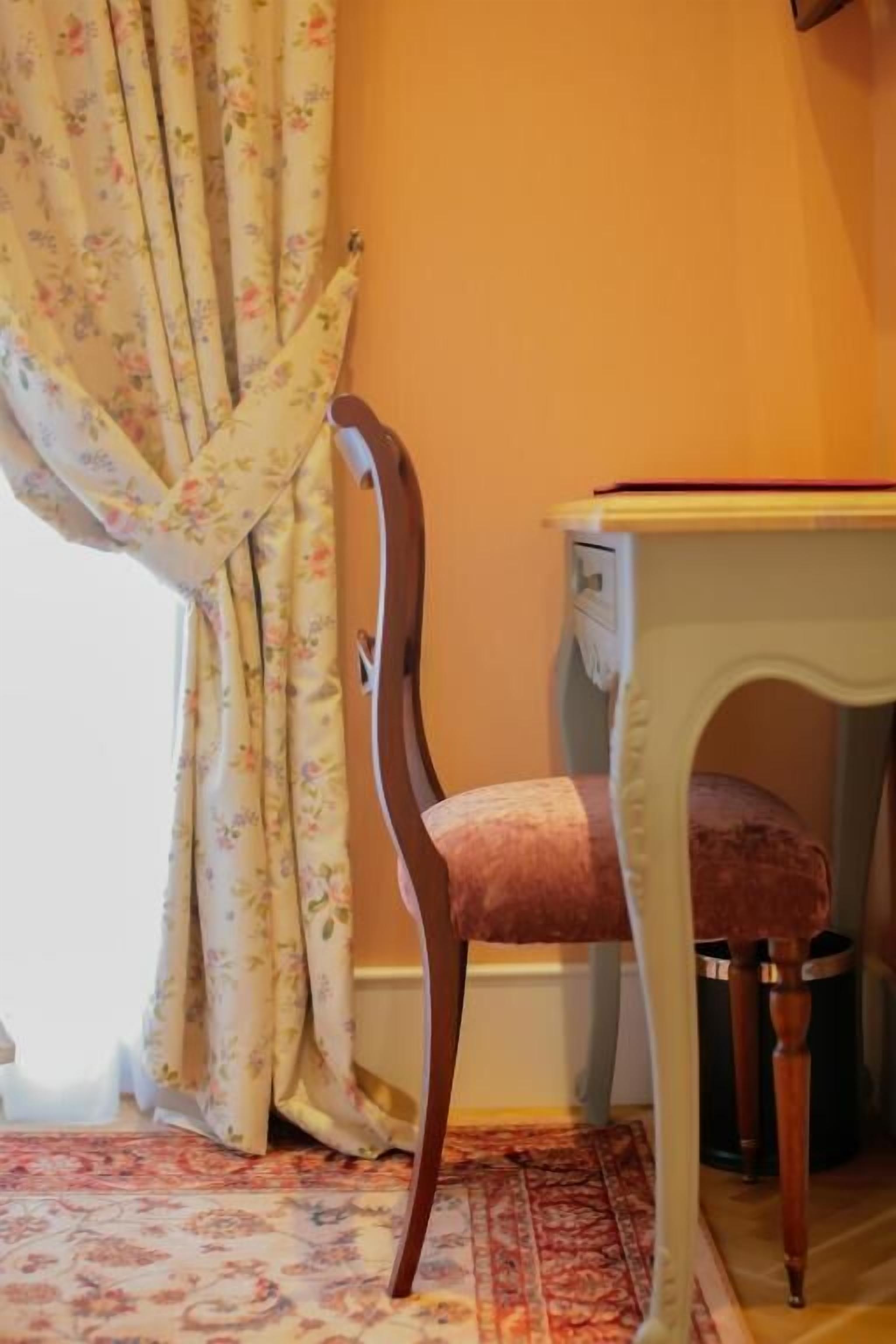 Una habitación con cortina de estampado floral, una silla de madera con cojín, una mesita blanca y un tapete estampado.