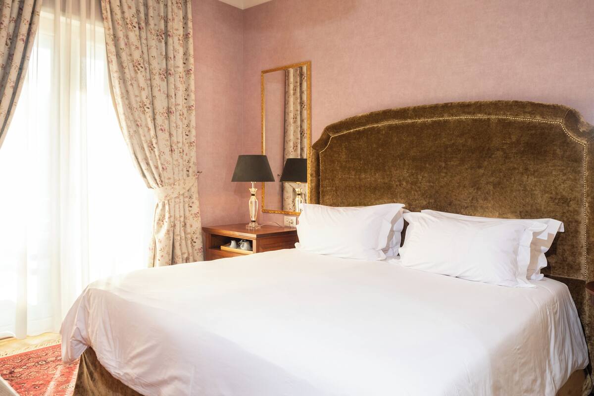A neatly made bed with white linens, a wooden nightstand with a lamp, a mirror, and a red patterned rug.