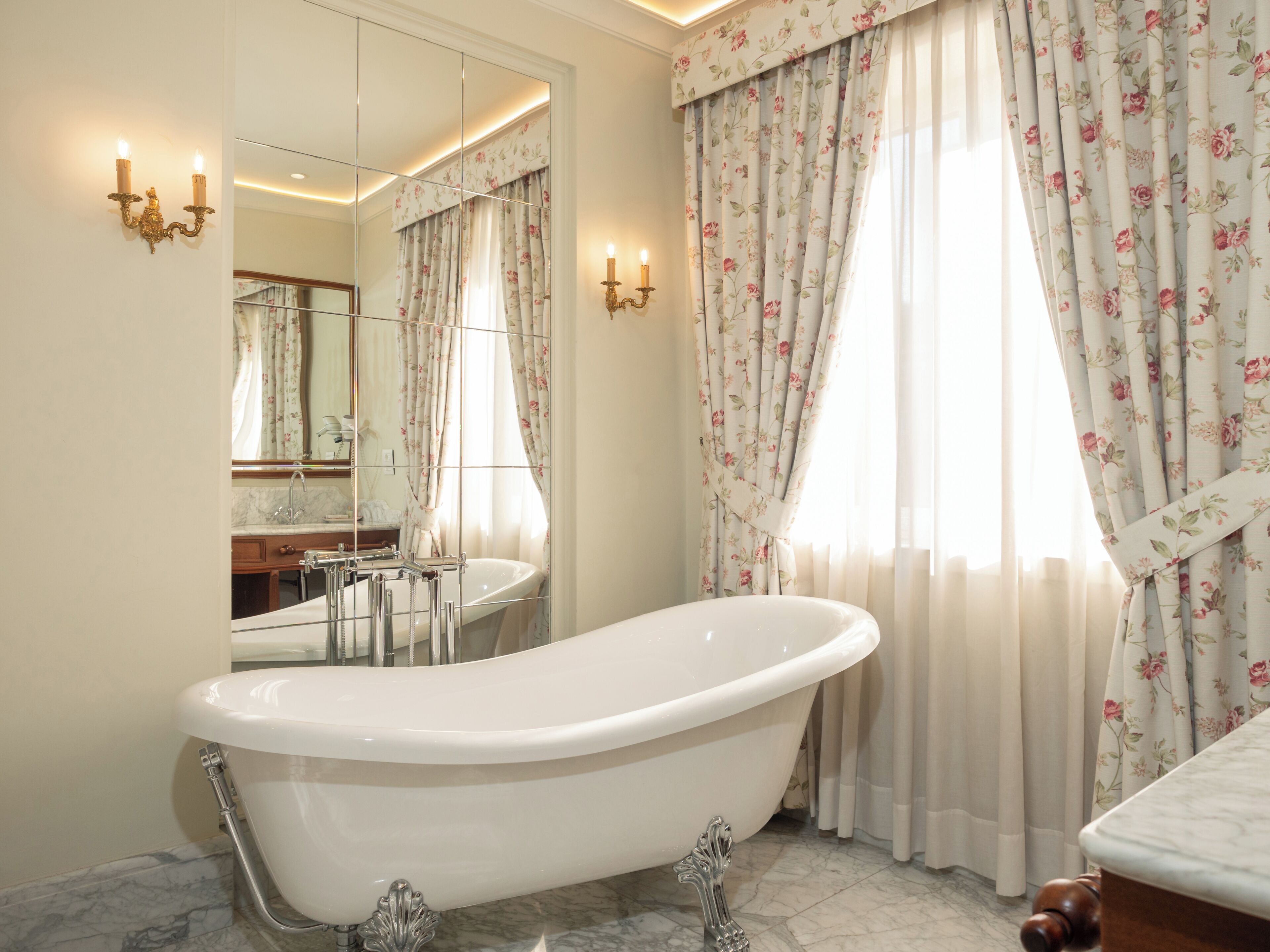 A bathroom with a freestanding bathtub, a mirror, a sink, and a window with floral curtains.