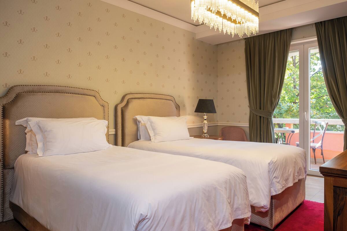 A hotel room with two beds, a chandelier, a balcony with a table and chairs, and a window with curtains.