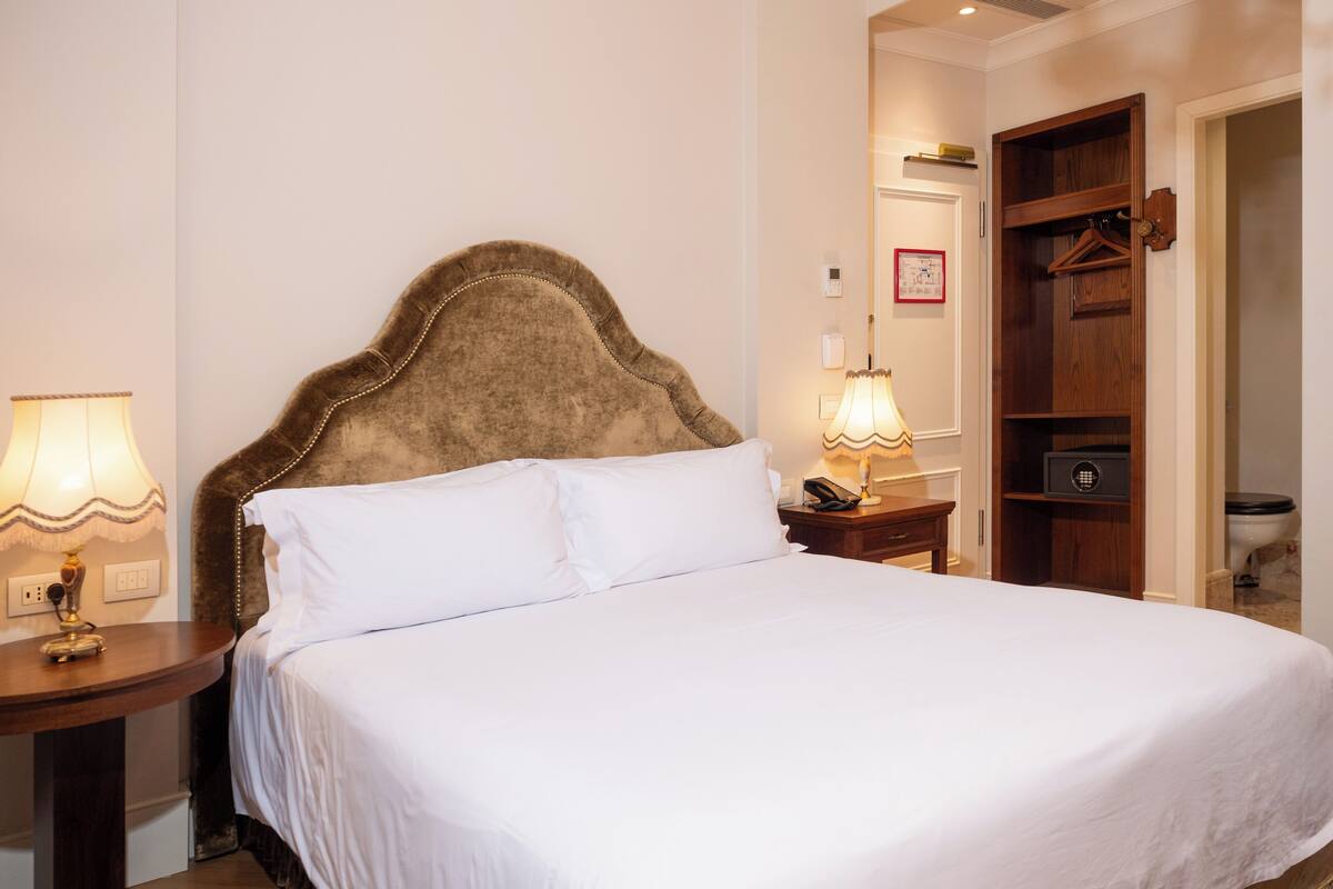 A neatly made bed with white linens, a wooden headboard, bedside lamps, and a wooden wardrobe with a hanging closet.