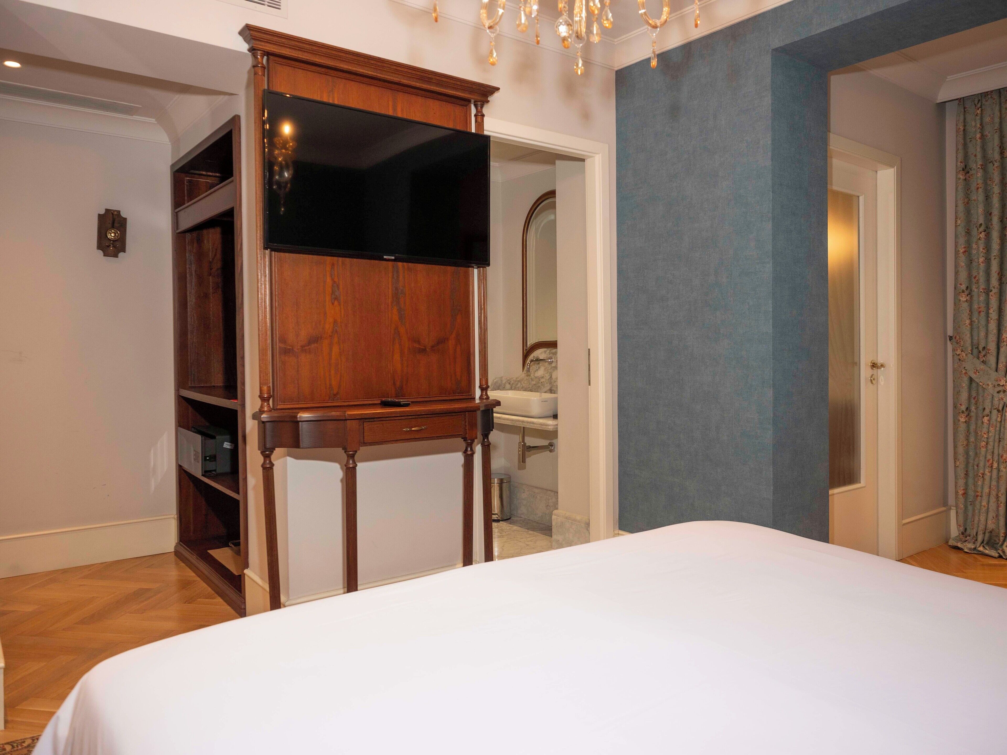 Un dormitorio con una cama grande, un mueble de televisión de madera, un baño visible a través de una puerta abierta y una araña.