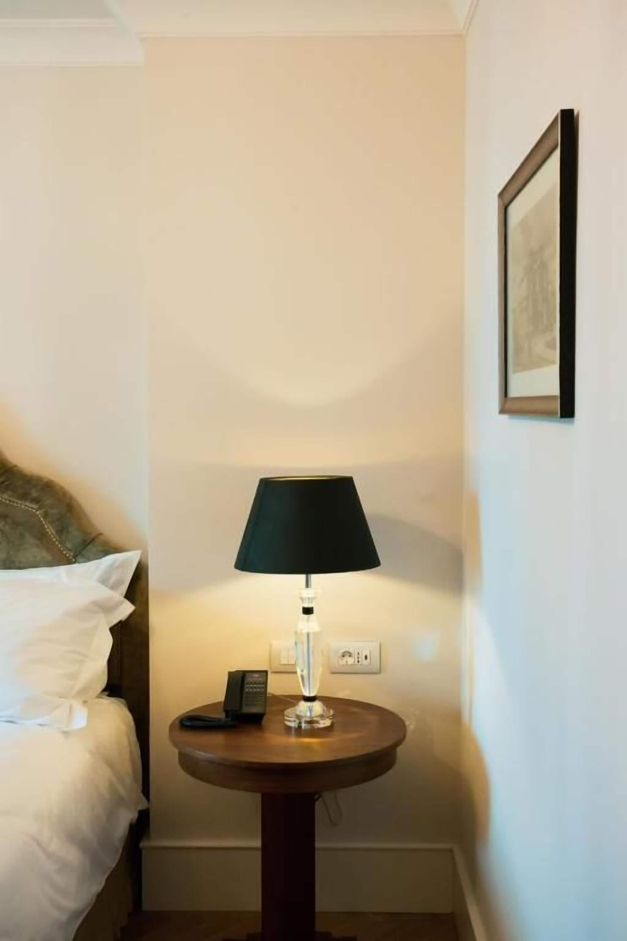 Una habitación de hotel con una cama, una mesa redonda con una lámpara, un teléfono y un cuadro en la pared.