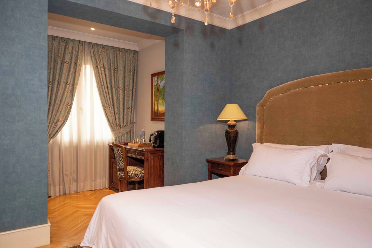 A hotel room with a large bed, a desk with a chair, a lamp, and a painting on the wall.