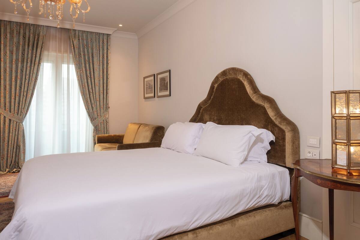 A bedroom with a large bed, a wooden nightstand, a chair, and a chandelier.