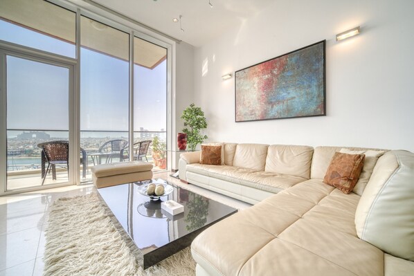 A spacious living room with a large sofa, a coffee table, and a view of the city through floor-to-ceiling windows. - Waterfront Sea View Apt w/ Wow Factor on The Palm (Dubai)
