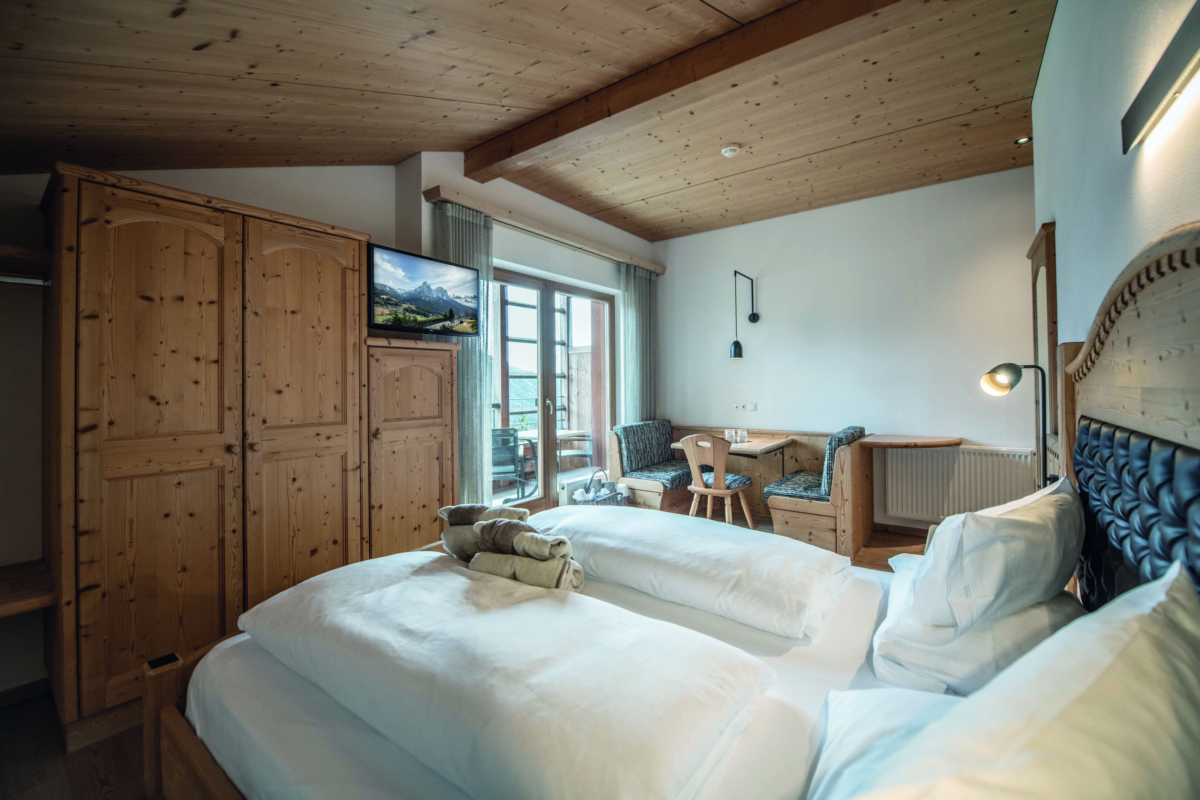 A bedroom with two beds, wooden wardrobe, desk, chair, and a view of the outdoors.