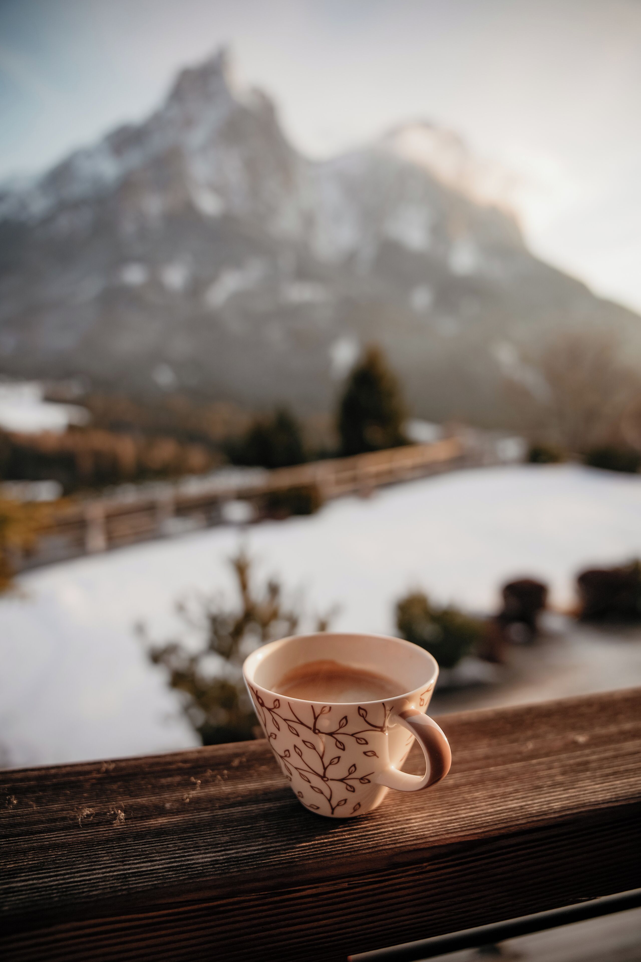 Una taza de café sobre un alféizar de madera, con un paisaje nevado y montañas al fondo.