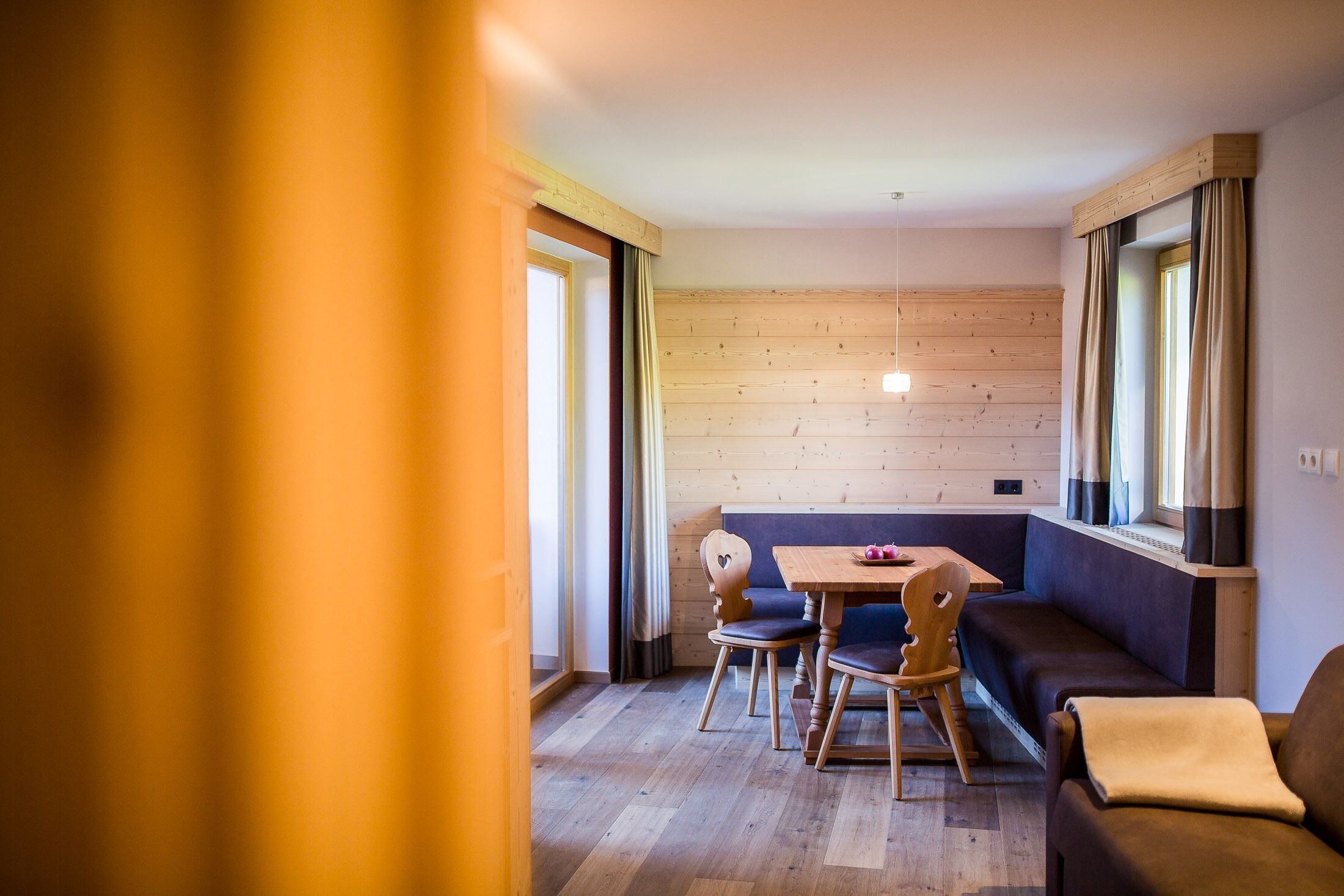 Una habitación acogedora con suelo de madera, un sofá, una mesa de comedor con sillas y ventanas grandes con cortinas.