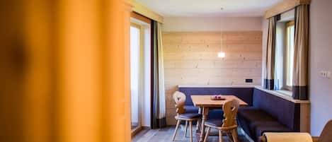 A cozy room with a wooden floor, a sofa, a dining table with chairs, and large windows with curtains.