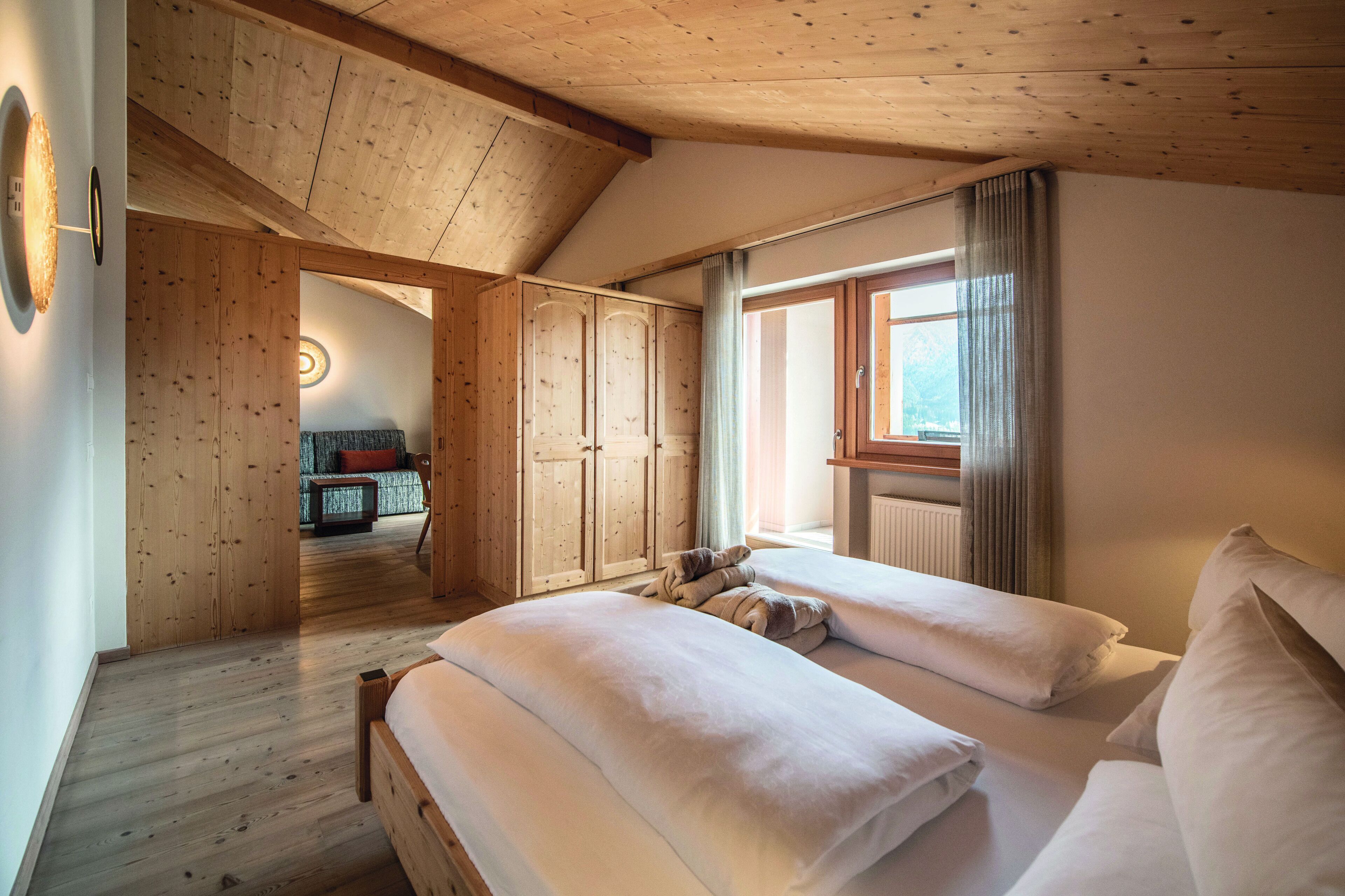 A bedroom with a wooden ceiling, a large window with curtains, and a bed with white bedding.