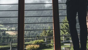 A person standing indoors, looking out at a mountainous landscape through a wooden-framed window. - Sonus Alpis - Adults Only (Castelrotto)