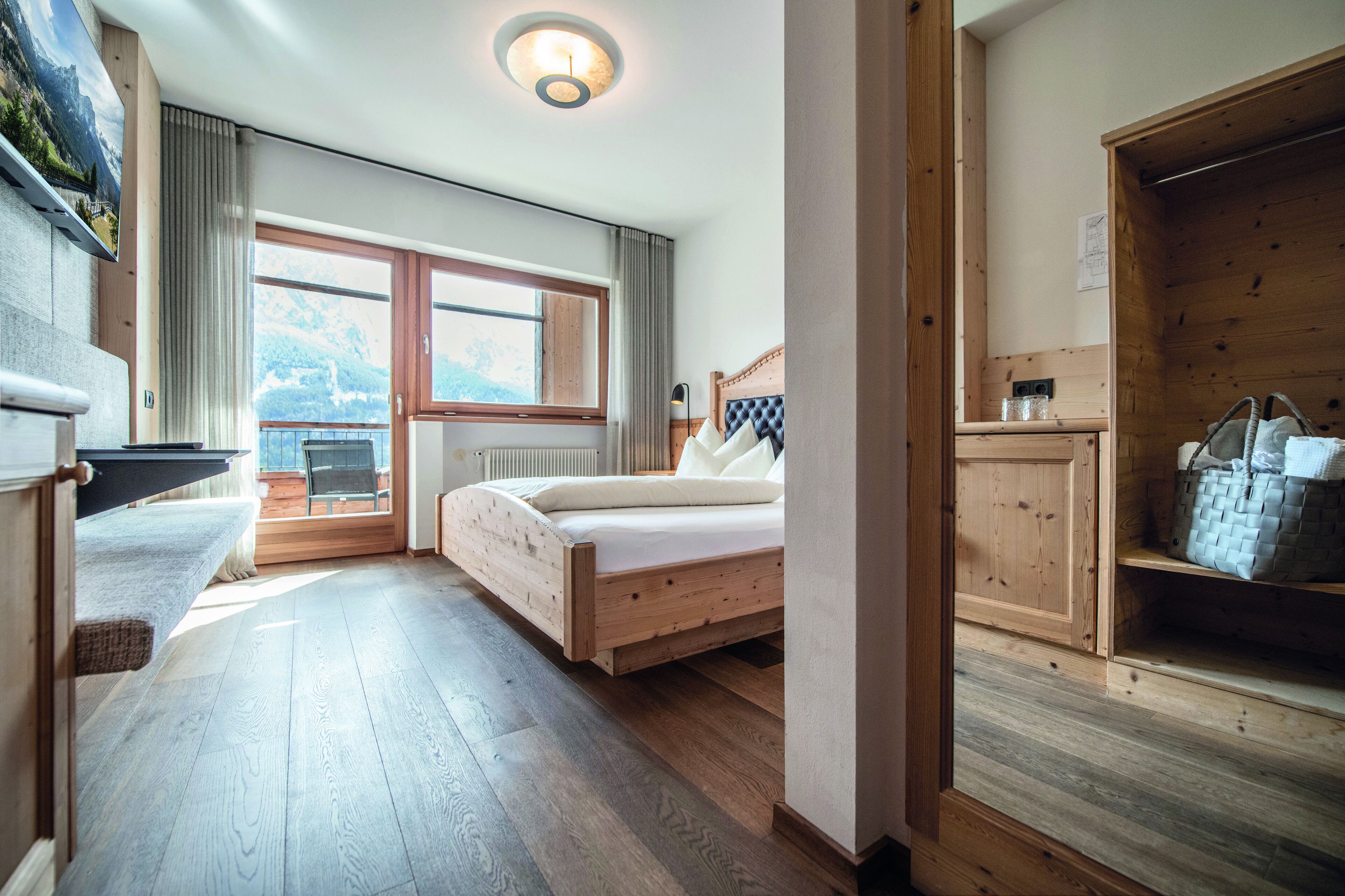 A bedroom with a wooden bed, a bench, a view of the outdoors, and a wooden wardrobe.