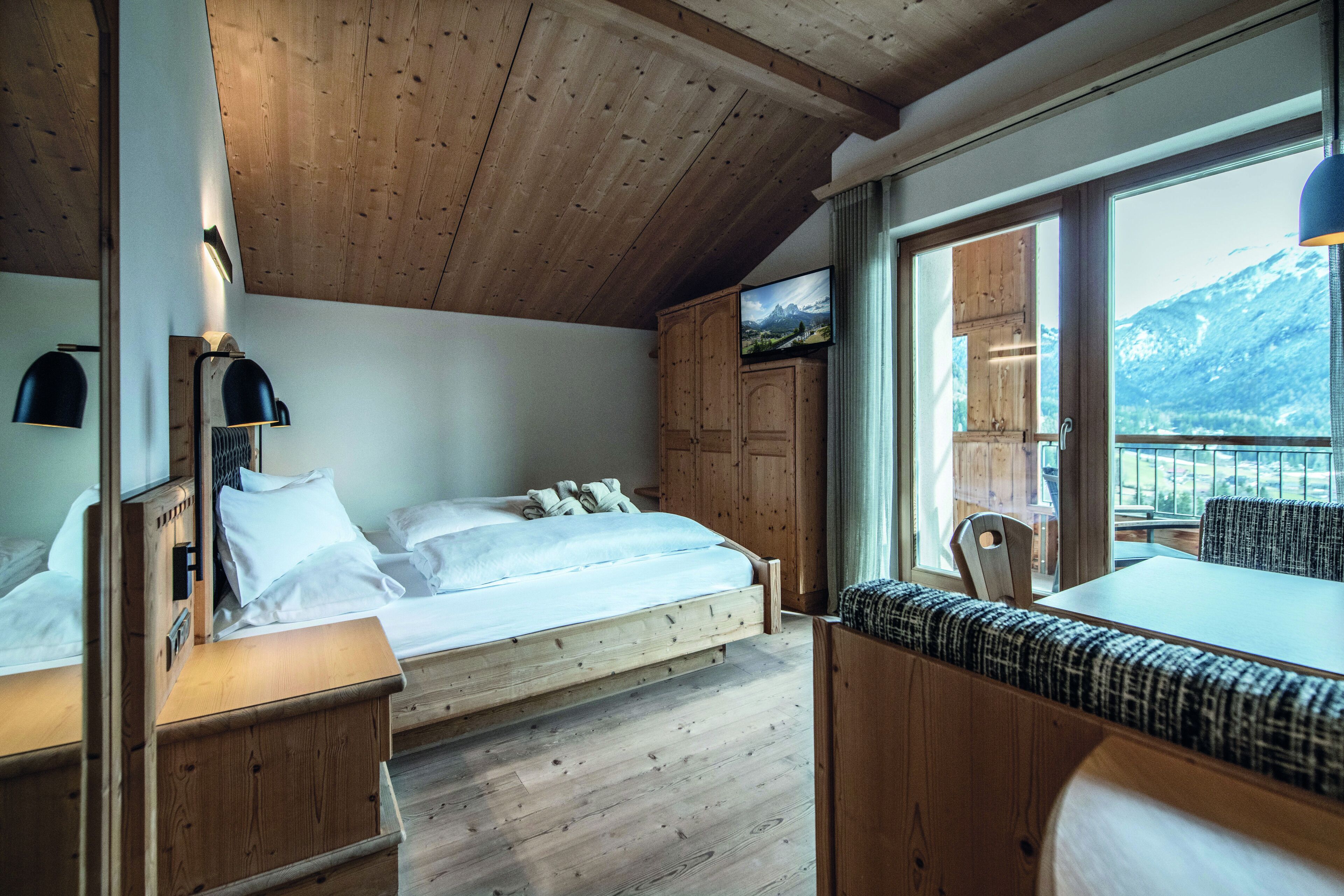 A bedroom with a wooden ceiling, a bed, a television, and a view of mountains through a window.