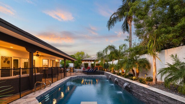 A swimming pool with a waterfall feature, surrounded by a patio area with outdoor seating and palm trees. - Pool, foosball, family fun at Always Summertime! (Scottsdale)