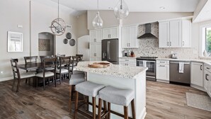 A modern kitchen with white cabinets, a central island with bar stools, a large refrigerator, and a dining area with wooden chairs and a table. - Pool, foosball, family fun at Always Summertime! (Scottsdale)