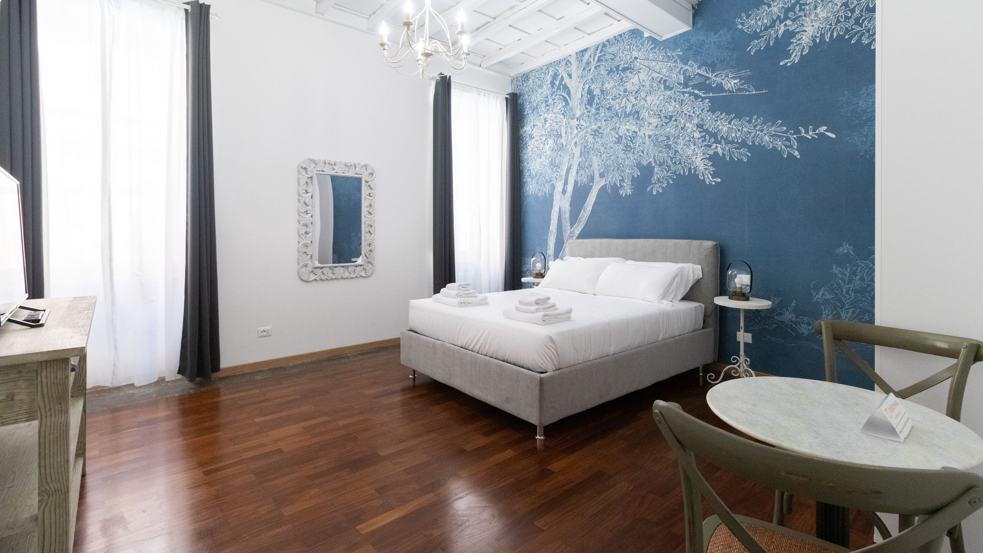 A bedroom with a bed, a bedside table, a chair, a mirror, and a wall with a tree mural.