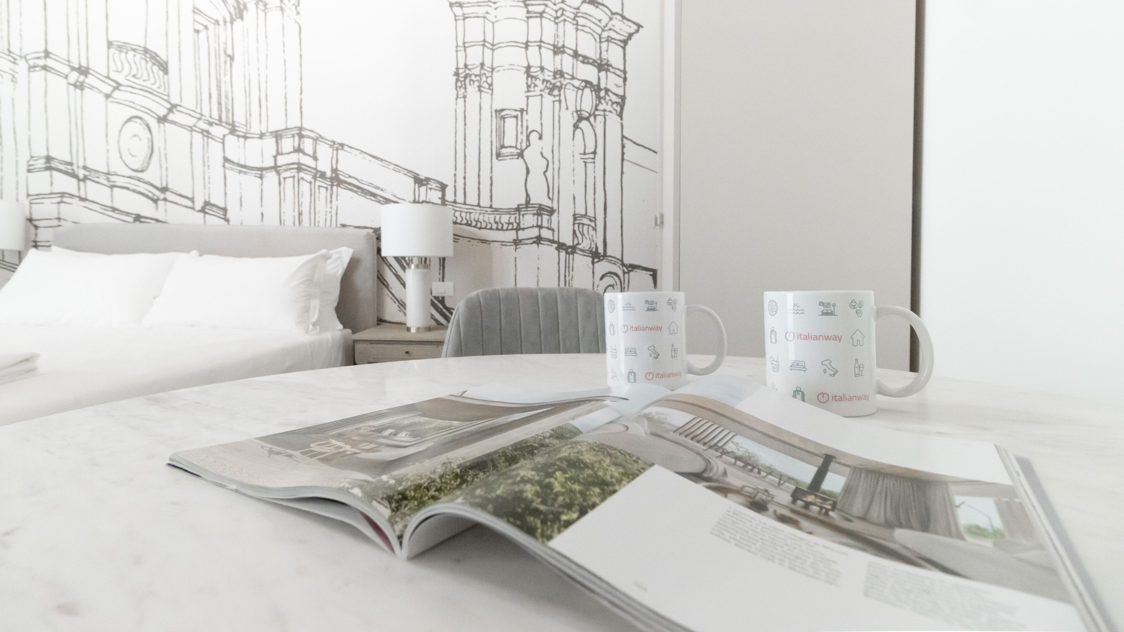 Una habitación de hotel con una cama, una mesita de noche, una lámpara, una silla y una mesa con una revista y dos tazas Italianway.