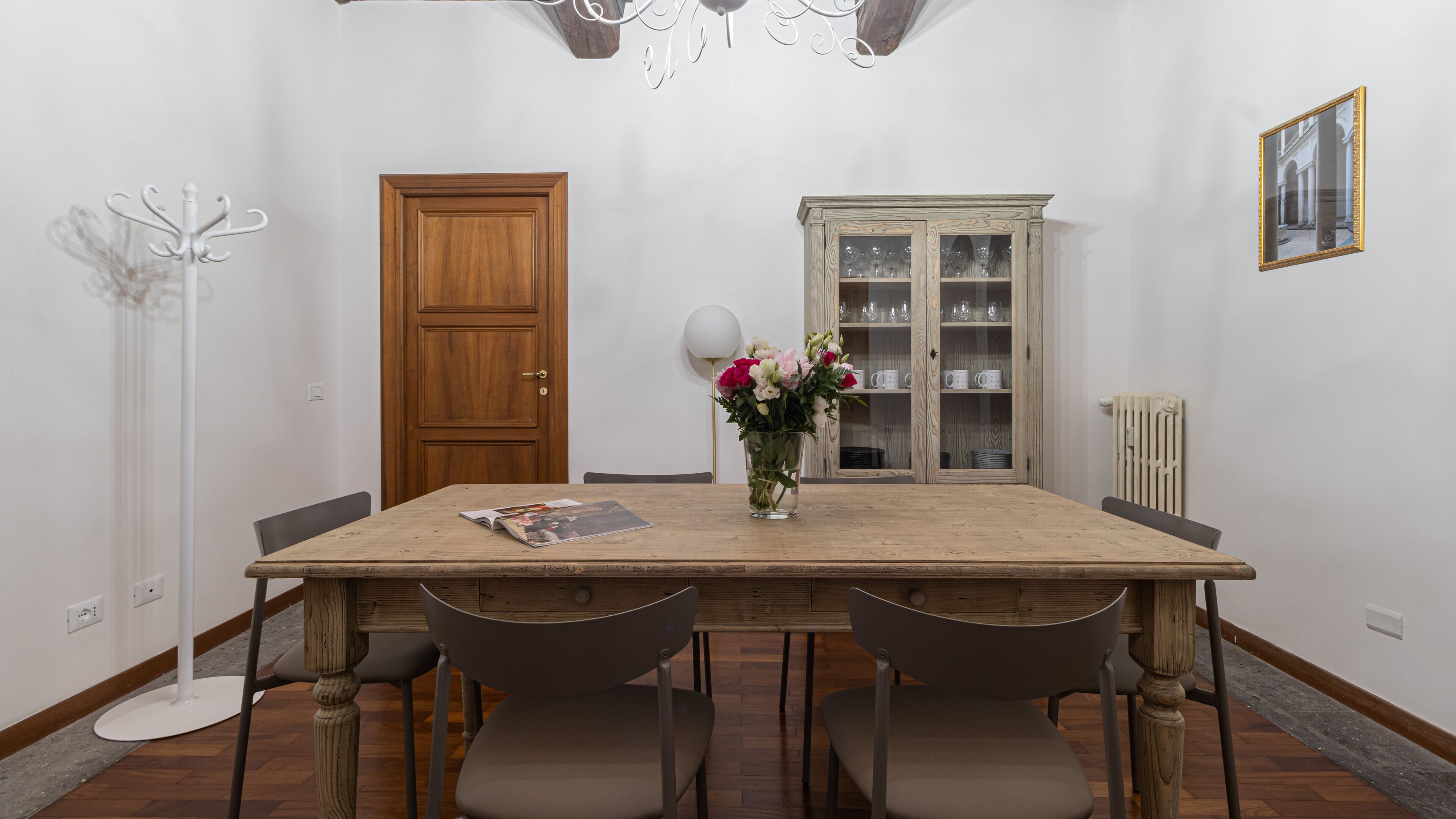 A dining area with a wooden table, four chairs, a vase with flowers, a cabinet with glass doors, and a coat rack.