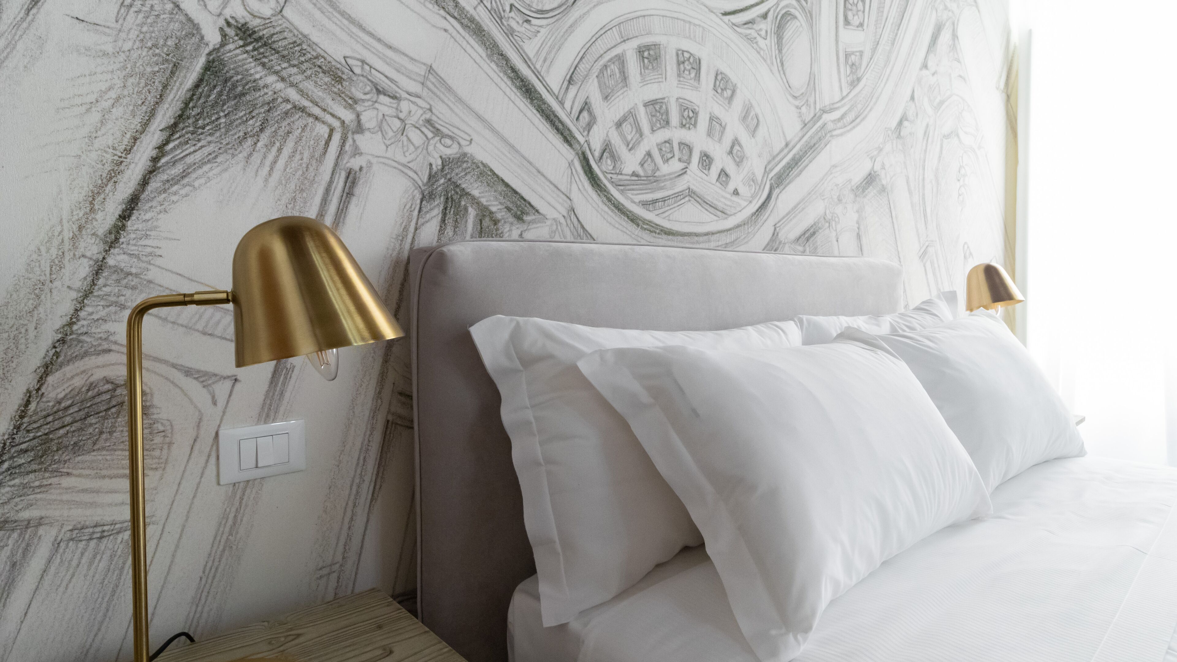 A hotel room with a bed, two bedside lamps, a wall with a detailed mural, and a power outlet.