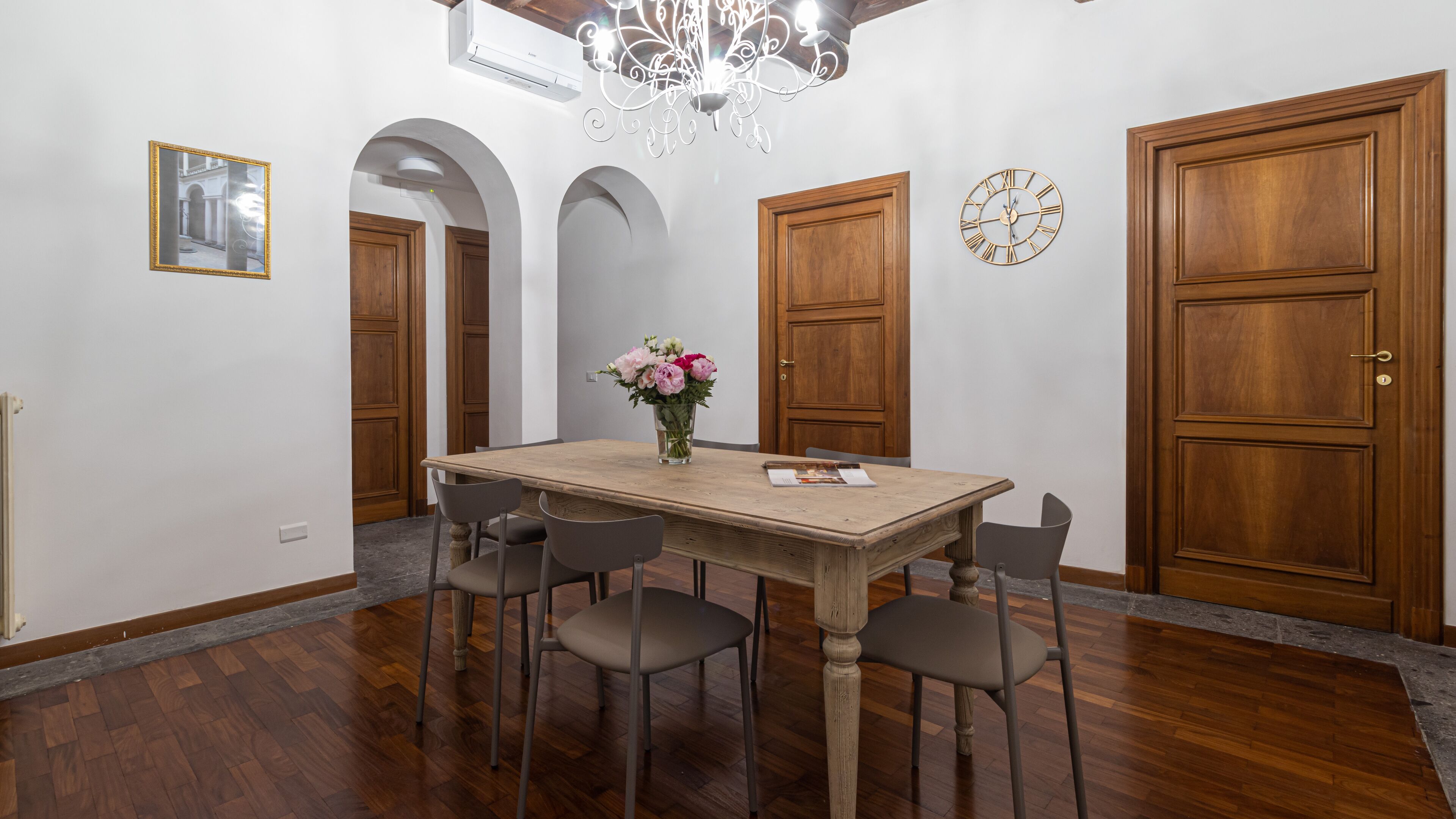 A dining area with a wooden table, four chairs, a vase with flowers, a clock, and wooden doors.